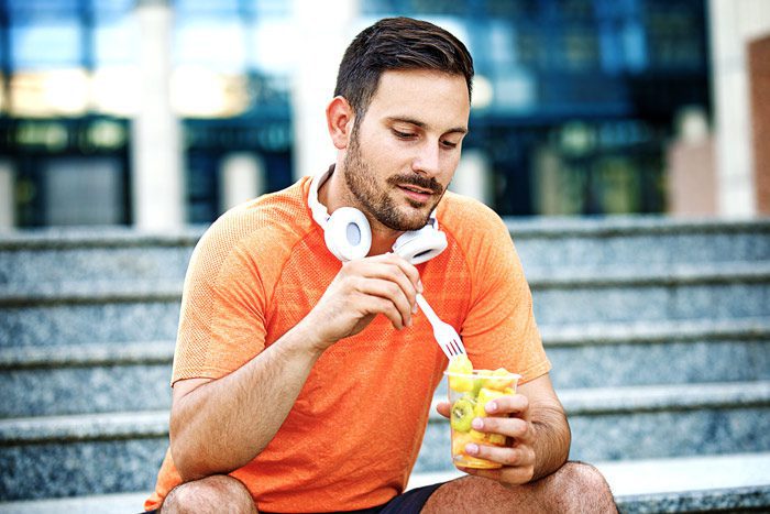 man in exercise clothes eating fruit salad outdoors on steps - self-care